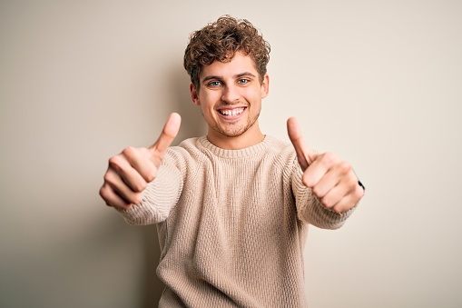 Young blond handsome man with curly hair wearing casual sweater over white background approving doing positive gesture with hand, thumbs up smiling and happy for success. Winner gesture.