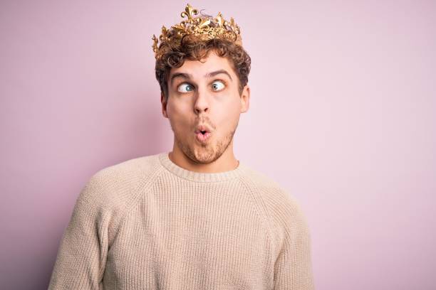 hombre rubio joven con el pelo rizado con corona dorada de rey sobre fondo rosa haciendo cara de pez con labios, gesto loco y cómico. expresión graciosa. - made man object fotografías e imágenes de stock