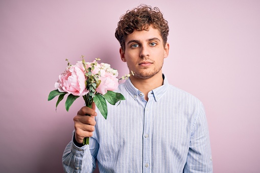 Young blond man with curly hair holding beautiful bouquet over isolated pink background with a confident expression on smart face thinking serious