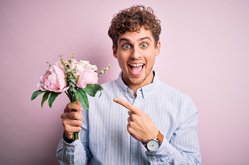 Young blond man with curly hair holding beautiful bouquet over isolated pink background very happy pointing with hand and finger