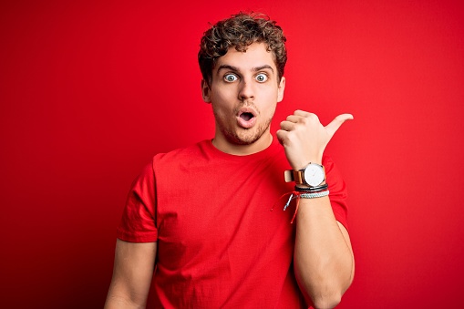 Young blond handsome man with curly hair wearing casual t-shirt over red background Surprised pointing with hand finger to the side, open mouth amazed expression.