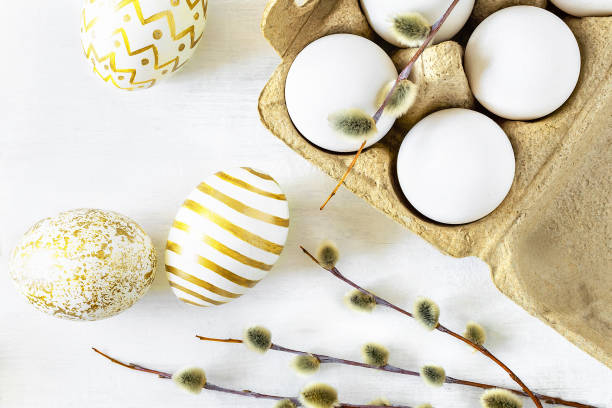 istock Easter unpainted chicken eggs in a cardboard box with a willow branch, some painted with gold paint patterns on a white table close-up. 1214284362
