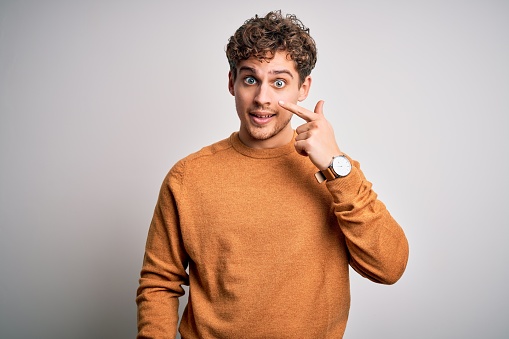 Young blond handsome man with curly hair wearing casual sweater over white background Pointing with hand finger to face and nose, smiling cheerful. Beauty concept