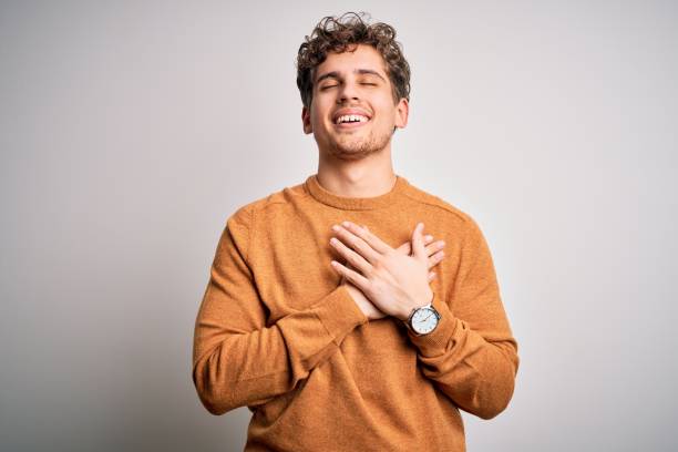 jeune homme beau blond avec les cheveux bouclés utilisant le chandail occasionnel au-dessus du fond blanc souriant avec des mains sur la poitrine avec les yeux fermés et le geste reconnaissant sur le visage. concept de santé. - torso male white men photos et images de collection