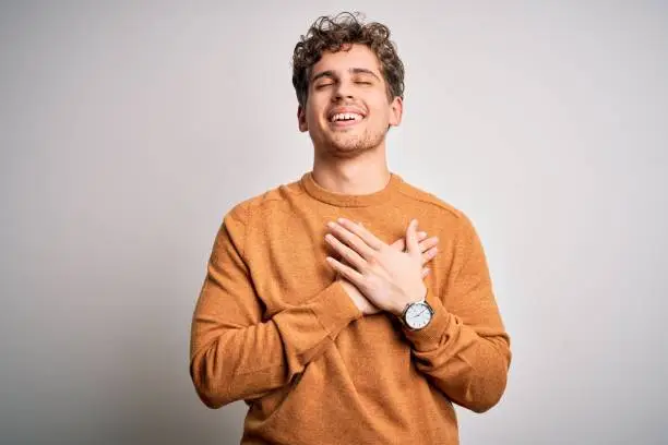 Young blond handsome man with curly hair wearing casual sweater over white background smiling with hands on chest with closed eyes and grateful gesture on face. Health concept.
