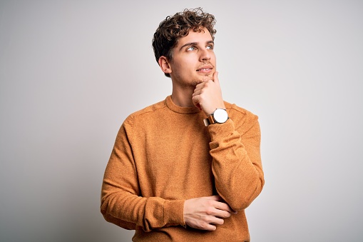 Young blond handsome man with curly hair wearing casual sweater over white background with hand on chin thinking about question, pensive expression. Smiling and thoughtful face. Doubt concept.