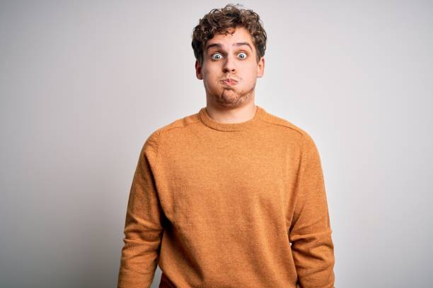 Young blond handsome man with curly hair wearing casual sweater over white background puffing cheeks with funny face. Mouth inflated with air, crazy expression. Young blond handsome man with curly hair wearing casual sweater over white background puffing cheeks with funny face. Mouth inflated with air, crazy expression. cheek stock pictures, royalty-free photos & images