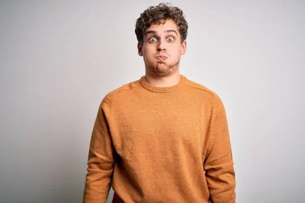 Young blond handsome man with curly hair wearing casual sweater over white background puffing cheeks with funny face. Mouth inflated with air, crazy expression.
