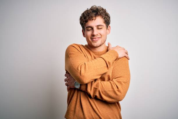 jeune homme beau blond avec les cheveux bouclés utilisant le chandail occasionnel au-dessus du fond blanc se serrant heureux et positif, souriant confiant. amour de soi et soins personnels - un seul homme photos et images de collection