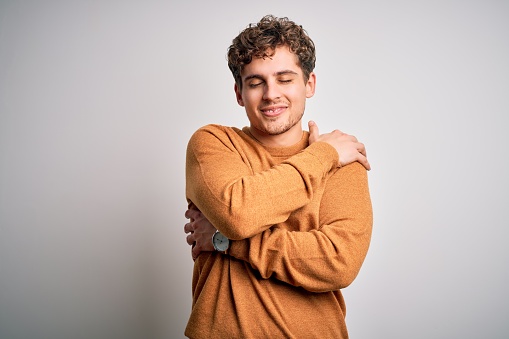 Young blond handsome man with curly hair wearing casual sweater over white background Hugging oneself happy and positive, smiling confident. Self love and self care