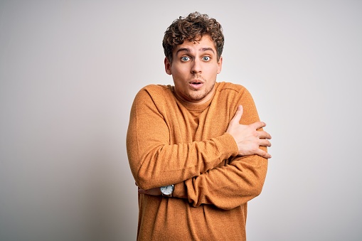 Young blond handsome man with curly hair wearing casual sweater over white background shaking and freezing for winter cold with sad and shock expression on face