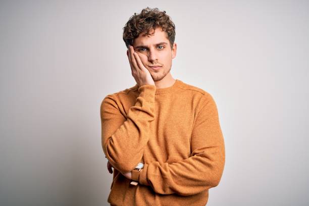 jeune homme blond beau avec les cheveux bouclés utilisant le chandail occasionnel au-dessus de la pensée blanche de fond regardant fatigué et ennuyé avec des problèmes de dépression avec les bras croisés. - dull colors photos et images de collection