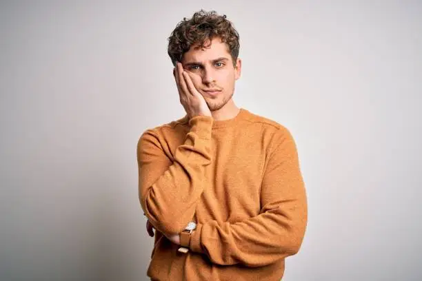 Photo of Young blond handsome man with curly hair wearing casual sweater over white background thinking looking tired and bored with depression problems with crossed arms.