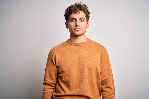 Young blond handsome man with curly hair wearing casual sweater over white background Relaxed with serious expression on face. Simple and natural looking at the camera.