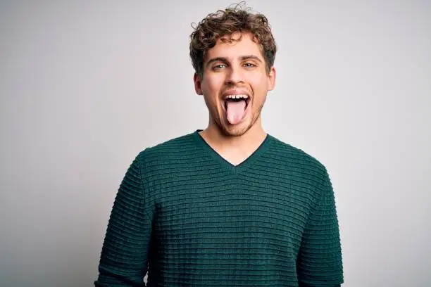 Young blond handsome man with curly hair wearing green sweater over white background sticking tongue out happy with funny expression. Emotion concept.