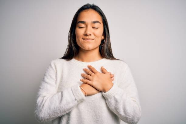 joven hermosa mujer asiática con suéter casual de pie sobre fondo blanco sonriendo con las manos en el pecho con los ojos cerrados y el gesto agradecido en la cara. concepto de salud. - white hands fotografías e imágenes de stock