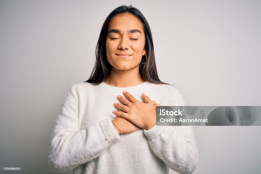 Young beautiful asian woman wearing casual sweater standing over white background smiling with hands on chest with closed eyes and grateful gesture on face. Health concept. Gratitude Stock Photo
