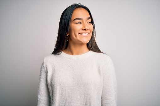 Young beautiful asian woman wearing casual sweater standing over white background looking away to side with smile on face, natural expression. Laughing confident.