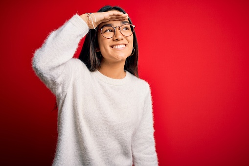 Young beautiful asian woman wearing casual sweater and glasses over red background very happy and smiling looking far away with hand over head. Searching concept.