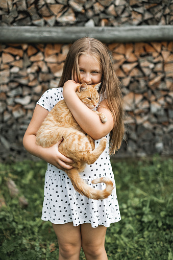 Close up portrait of beautiful cute girl with long hair wearing white dress, smiling, holding big red cat on hands, and looking at the camera, standing over firewood background, summer holidays.