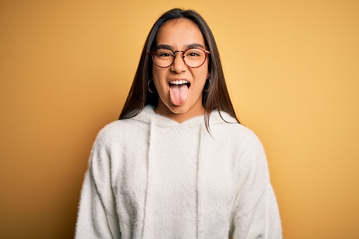 Young beautiful asian woman wearing casual sweater and glasses over yellow background sticking tongue out happy with funny expression. Emotion concept.