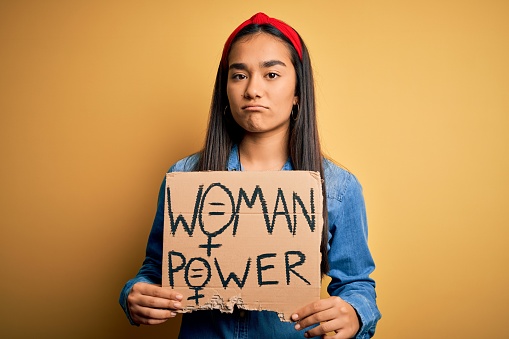 Beautiful activist asian woman asking for women rights holding banner with power message with a confident expression on smart face thinking serious