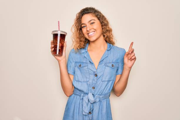 Beautiful blonde woman with blue eyes drinking cola beverage using straw to refreshment surprised with an idea or question pointing finger with happy face, number one