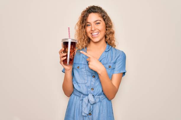 istock Beautiful blonde woman with blue eyes drinking cola beverage using straw to refreshment very happy pointing with hand and finger 1214302316