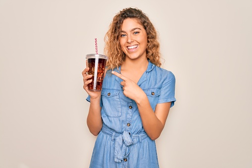 Beautiful blonde woman with blue eyes drinking cola beverage using straw to refreshment very happy pointing with hand and finger