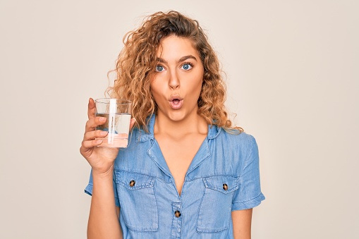 Young beautiful blonde woman with blue eyes drinking glass of water over white background scared in shock with a surprise face, afraid and excited with fear expression