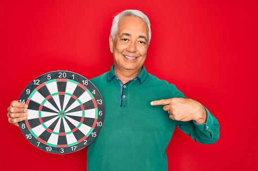 Middle age senior grey-haired man holding competition dartboard target over red background with surprise face pointing finger to himself