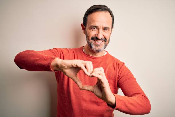 hombre canoso de la edad media con suéter naranja casual de pie sobre fondo blanco aislado sonriendo en el amor haciendo forma de símbolo del corazón con las manos. concepto romántico. - made man object fotografías e imágenes de stock