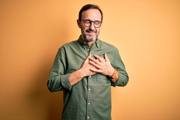 middle age hoary man wearing casual green shirt and glasses over isolated yellow background smiling with hands on chest with closed eyes and grateful gesture on face. health concept. - peito imagens e fotografias de stock