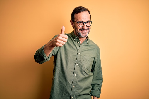 Middle age hoary man wearing casual green shirt and glasses over isolated yellow background doing happy thumbs up gesture with hand. Approving expression looking at the camera showing success.