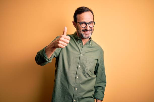 Middle age hoary man wearing casual green shirt and glasses over isolated yellow background doing happy thumbs up gesture with hand. Approving expression looking at the camera showing success. Middle age hoary man wearing casual green shirt and glasses over isolated yellow background doing happy thumbs up gesture with hand. Approving expression looking at the camera showing success. thumb stock pictures, royalty-free photos & images