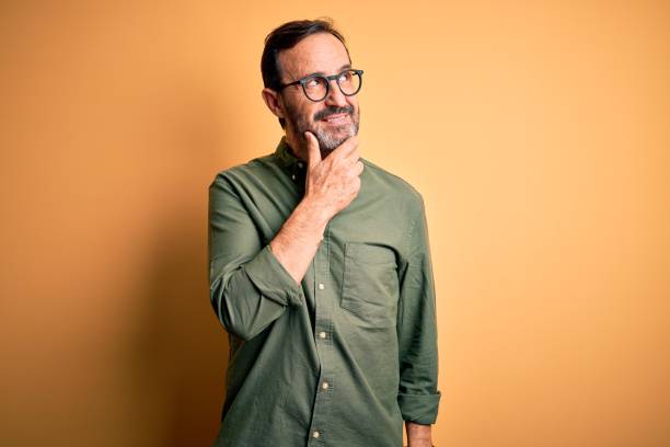 Middle age hoary man wearing casual green shirt and glasses over isolated yellow background with hand on chin thinking about question, pensive expression. Smiling with thoughtful face. Doubt concept. Middle age hoary man wearing casual green shirt and glasses over isolated yellow background with hand on chin thinking about question, pensive expression. Smiling with thoughtful face. Doubt concept. man thinking stock pictures, royalty-free photos & images