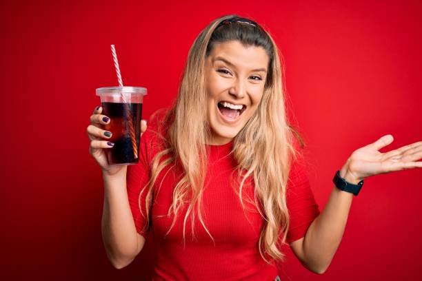 istock Young beautiful blonde woman drinking cola fizzy beverage using straw over red background very happy and excited, winner expression celebrating victory screaming with big smile and raised hands 1214317080