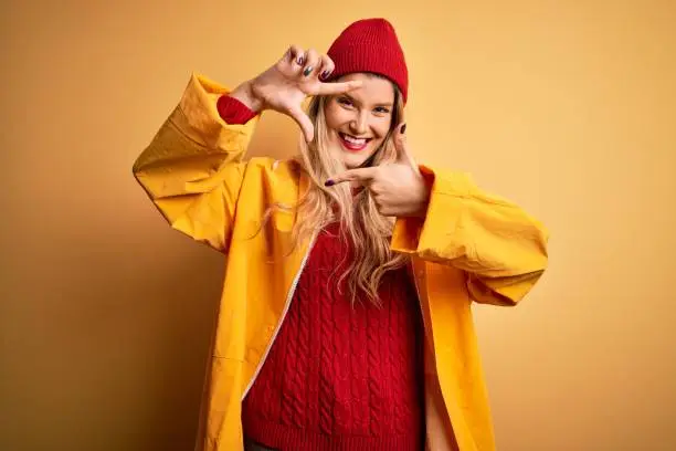 Photo of Young beautiful blonde woman wearing raincoat and wool cap over isolated yellow background smiling making frame with hands and fingers with happy face. Creativity and photography concept.