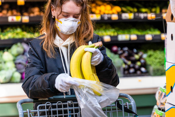 pandemic times shopping. a young woman wearing a protective mask and gloves buying babanas in a supermarket. - healthy eating healthcare and medicine healthy lifestyle people imagens e fotografias de stock