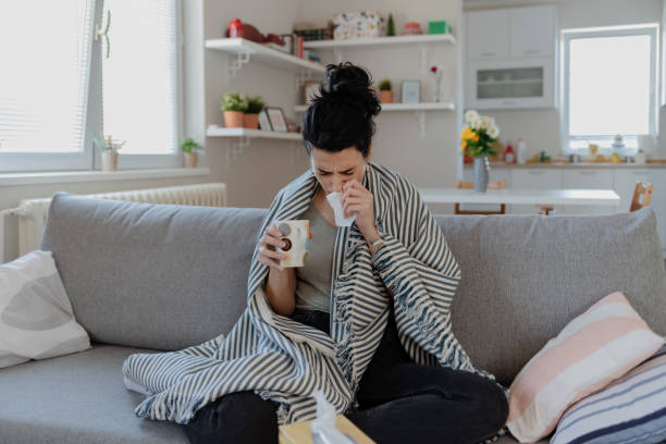 Sick Woman Sick Woman Covered With a Blanket Lying in Bed With High Fever and a Flu, Resting at Living Room. She Is Exhausted and Suffering From Flu. Sick Woman With Runny Nose Lying in Bed. Girl Suffering From Cold Lying in Bed With Tissue Blowing Her Nose While Sitting on the Sofa symptom stock pictures, royalty-free photos & images