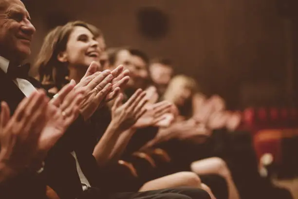 Side view of smiling spectators clapping hands in opera house. Men and women are watching theatrical performance. They are in elegant wear.