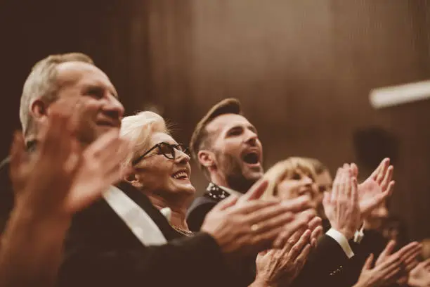 Smiling audience clapping in the opera. Focus on man shouting. Men and women are watching theatrical performance. They are in elegant wear.