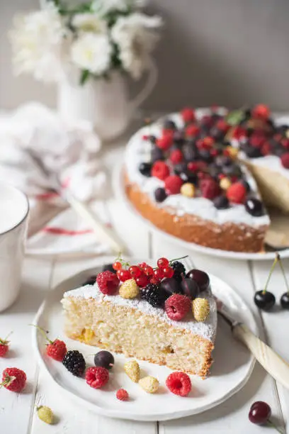 Homemade sponge cake with a variety of berries on a white plate on a white wooden background