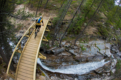 A male cyclist crosses a bridge over a stream during a mountain bike ride on the High Rockies Trail in Alberta, Canada. The High Rockies Trail is part of The Great Trail or the Trans Canada Trail which spans the width of Canada. He is riding a modern enduro-style mountain bike, wears a helmet and carries a hydration backpack.