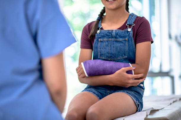 Young Girl with Broken Arm Visits Doctor stock photo A young multi-ethnic girl with a purple cast on her arm visits an Asian female doctor.  The doctor is standing with her back to the camera and the young girl sits on the examination table. orthopedic cast stock pictures, royalty-free photos & images