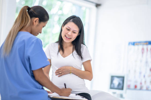 Pregnant Woman Visits Female Doctor stock photo A pregnant Asian woman sits with her hands on her belly as she visits a mixed race female doctor.  The doctor is wearing blue scrubs and has her back to the camera. cambodian ethnicity stock pictures, royalty-free photos & images