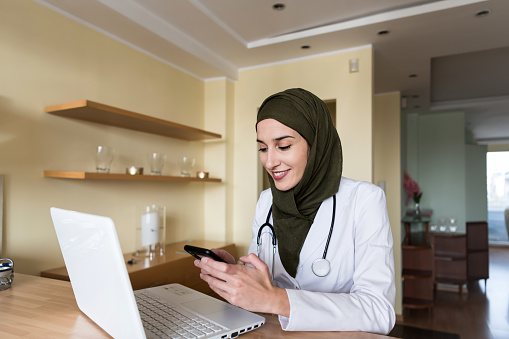 A Muslim female doctor is sitting at a work desk at home and typing messages on her mobile phone. A happy and confident medicine worker of Arabian ethnicity is using her laptop in her domestic room.