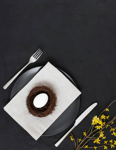 istock Easter minimal table setting with a chicken egg in a nest and yellow flowers on a black background. Vertical orientation. 1214378497