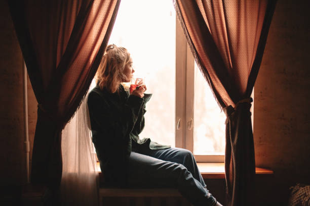 istock Young woman drinking tea sitting on windowsill at home 1214387147