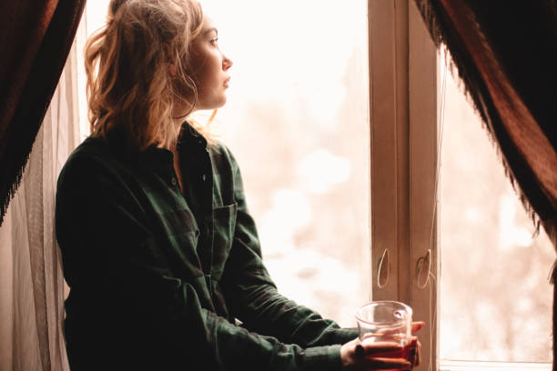 istock Young woman drinking tea sitting on windowsill at home 1214387338